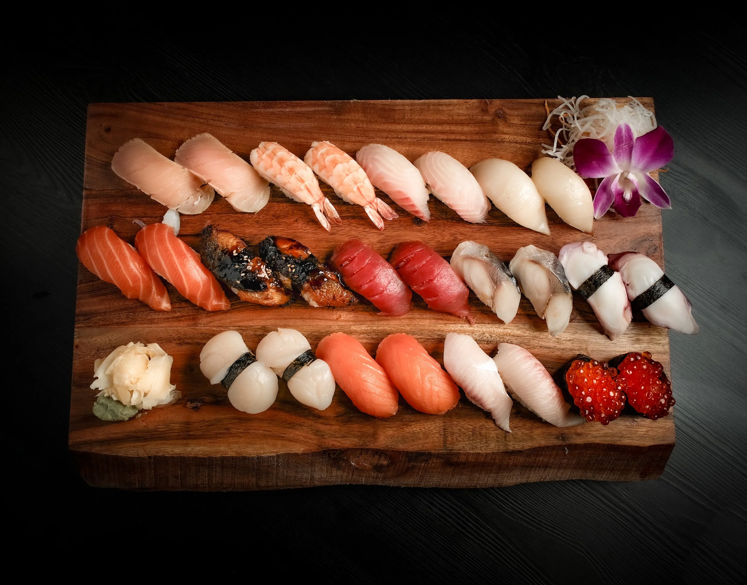 Assorted sushi on a wooden platter, including various types of raw fish, shrimp, and sushi rolls, garnished with shredded daikon and a purple orchid flower.