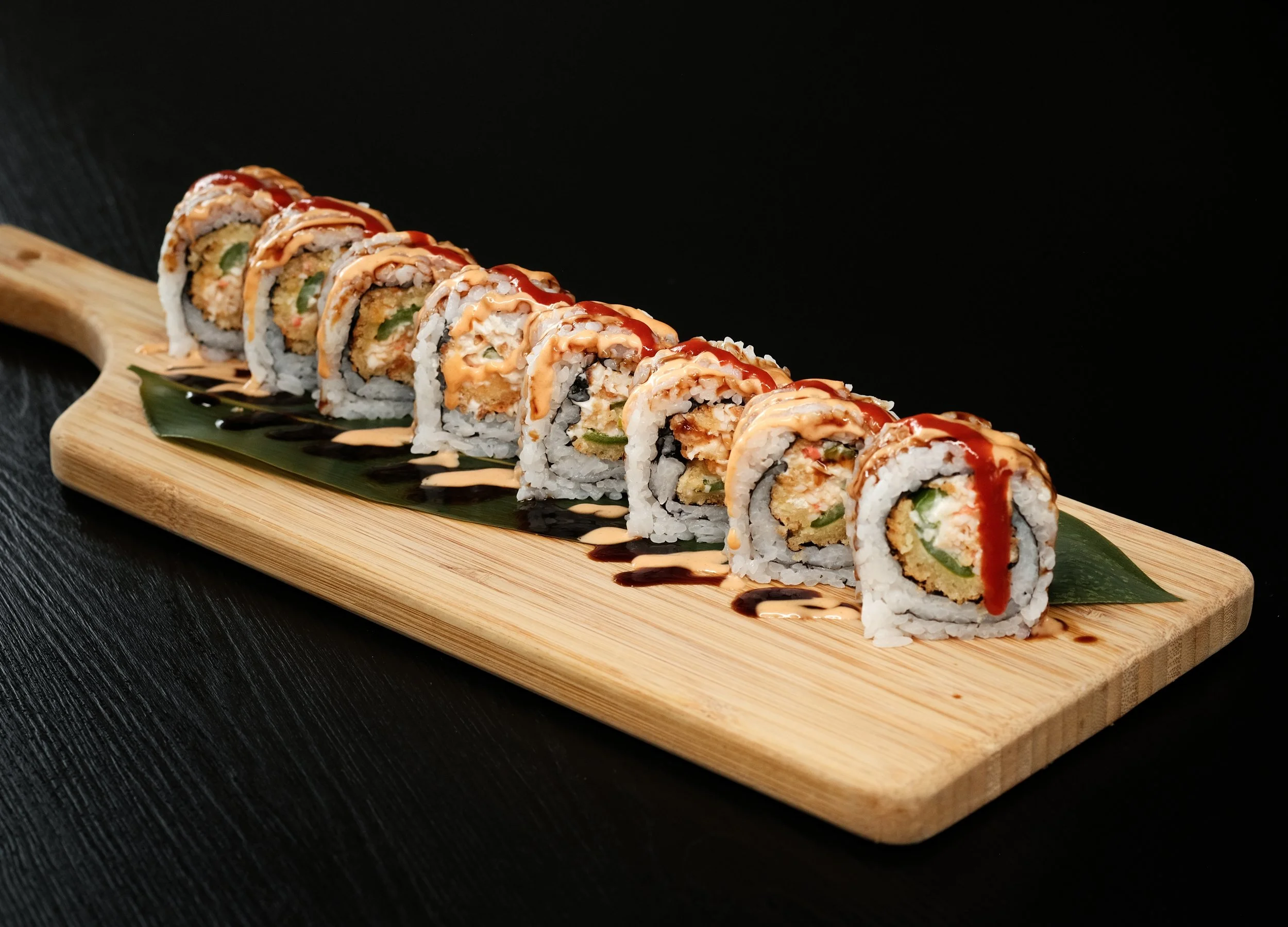 Sushi rolls drizzled with sauces on a wooden platter with a leaf garnish against a black background