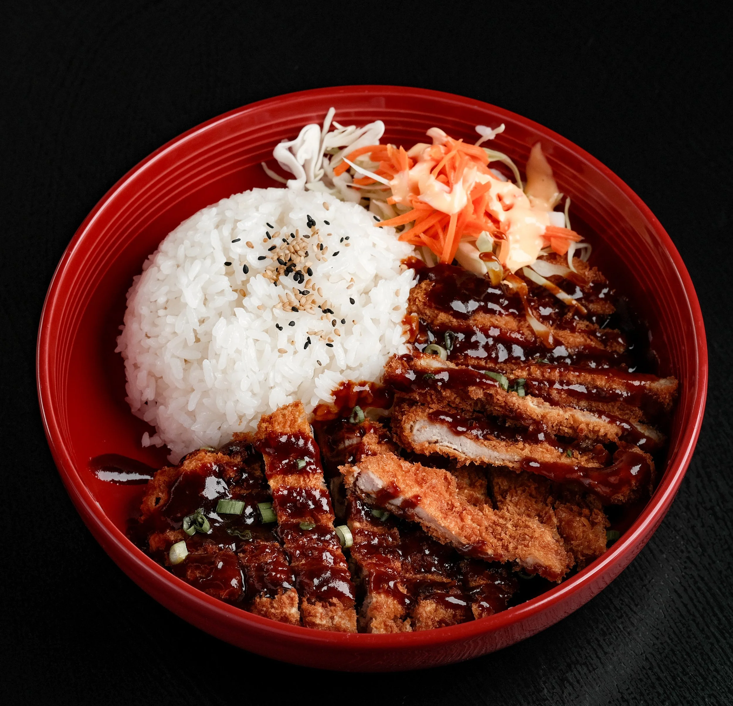A red bowl contains white rice topped with black sesame seeds, serving as a base for breaded and fried meat cutlets with dark sauce, accompanied by shredded cabbage and coleslaw with dressing.