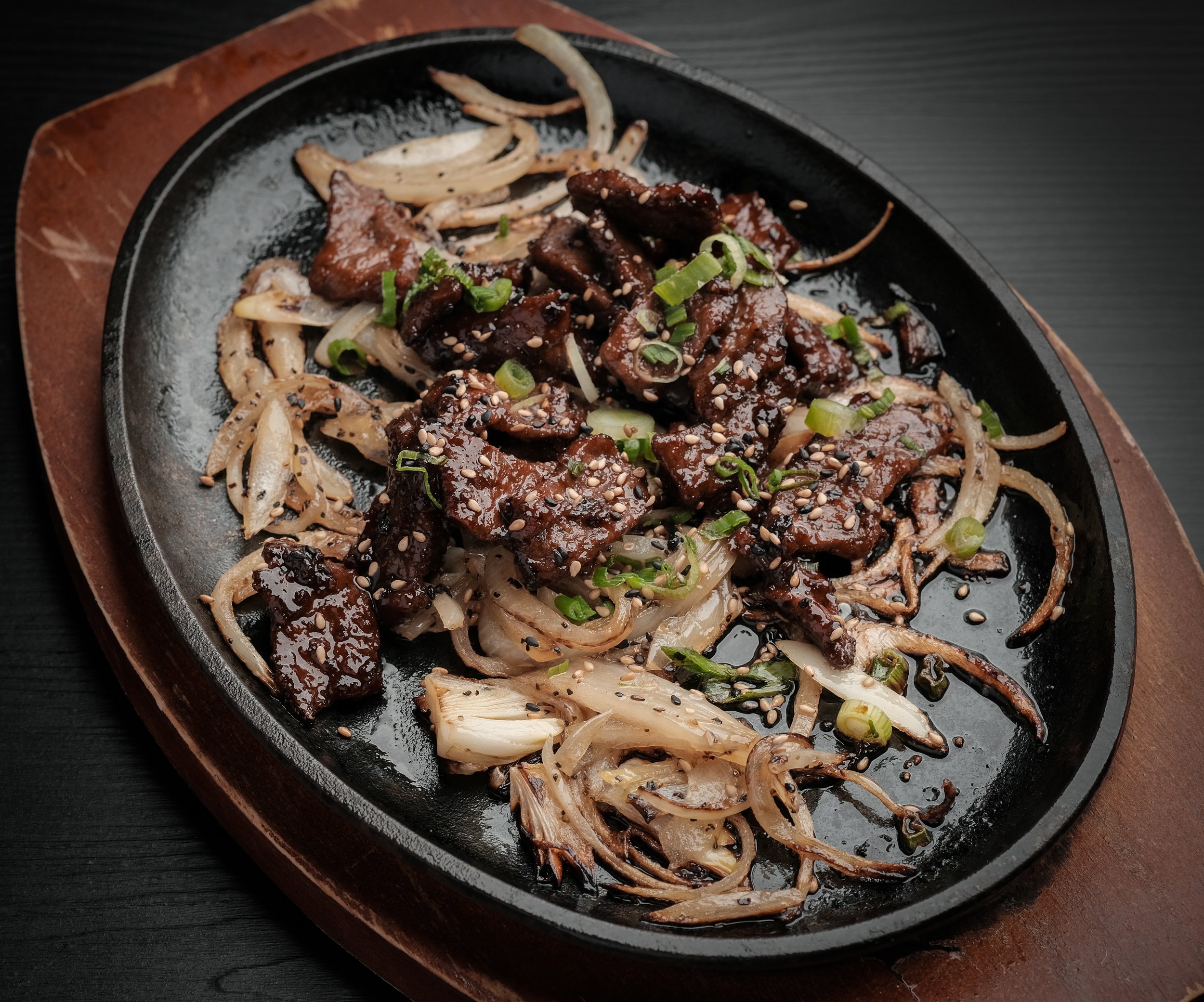 Sweet and savory grilled beef served on a sizzling cast iron skillet, garnished with chopped green onions and sprinkled with sesame seeds, accompanied by caramelized onions and cooked shredded vegetables.