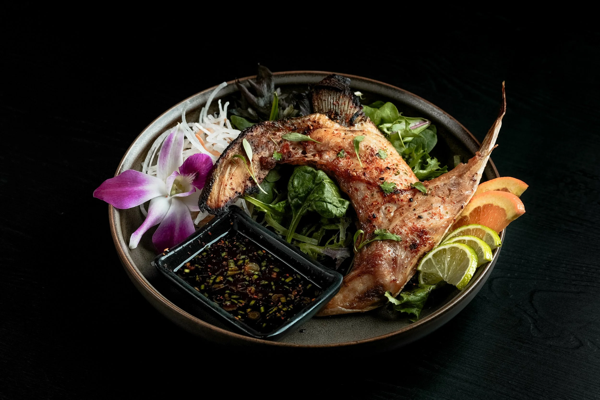 Grilled fish served on a bed of mixed greens with lemon and lime slices, orchid and other edible flowers, and a small dish of dipping sauce.