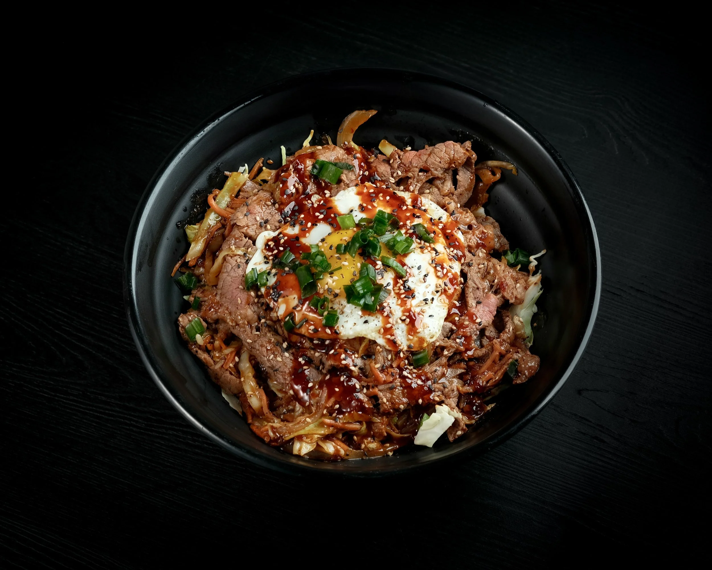 Bowl of Korean-style beef with vegetables, topped with a fried egg, chopped green onions, sesame seeds, and sauce, on a black wooden surface.
