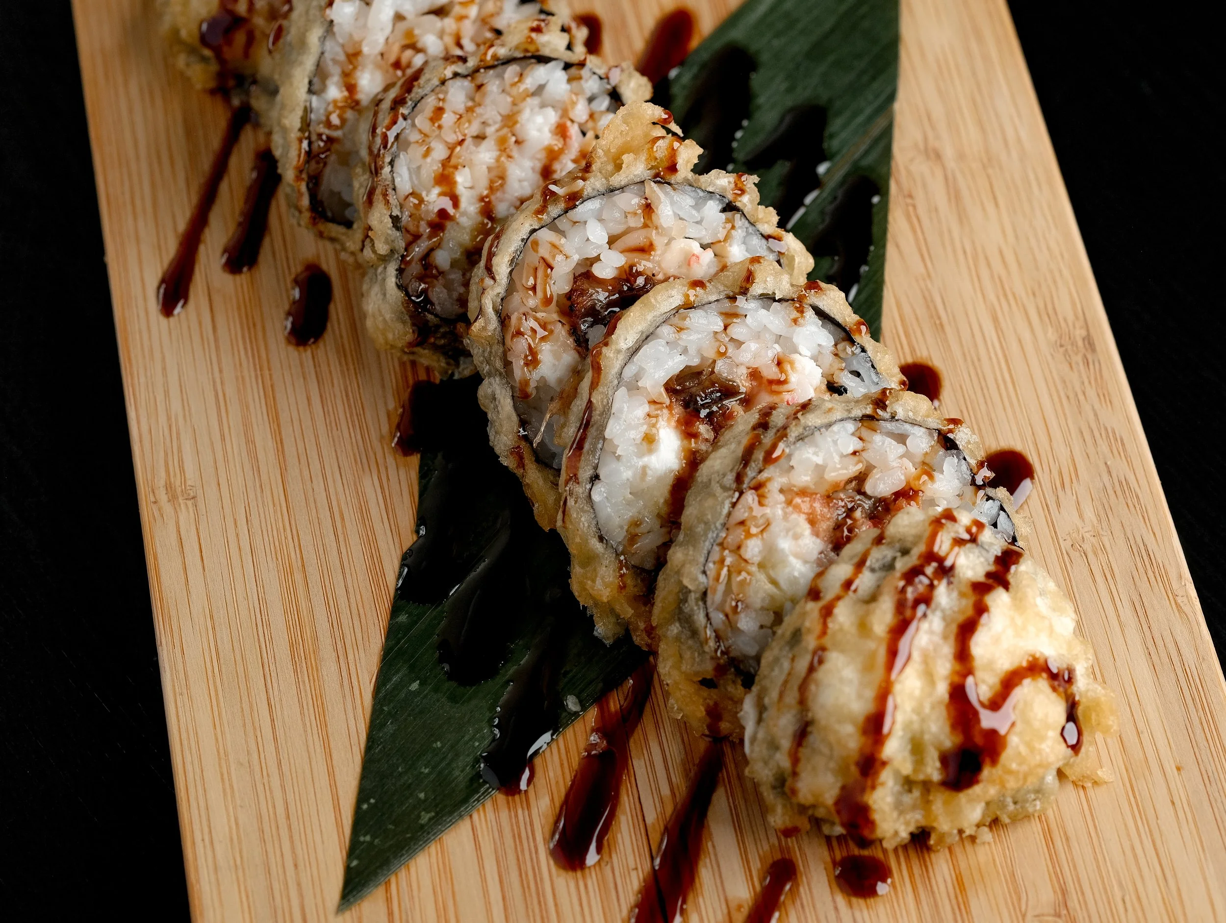 A plate of sushi rolls with tempura, topped with drizzled eel sauce and served on a bamboo board with a green leaf garnish.