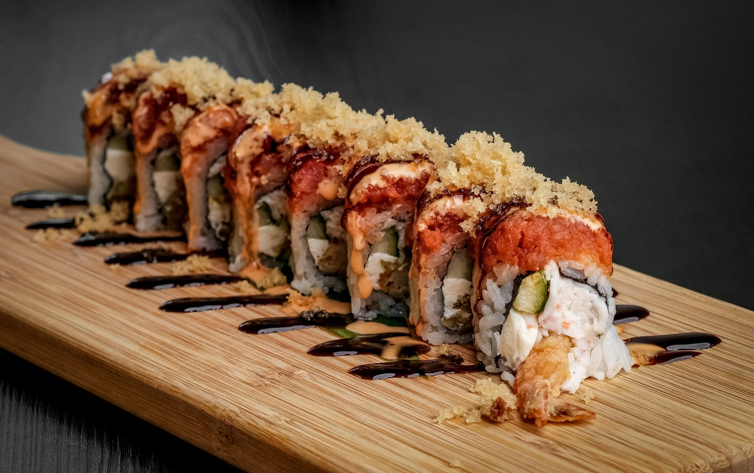 Close-up of a sushi roll with tempura shrimp, cucumber, cream cheese, topped with spicy mayo, eel sauce, and crispy tempura flakes, served on a wooden platter with black background.