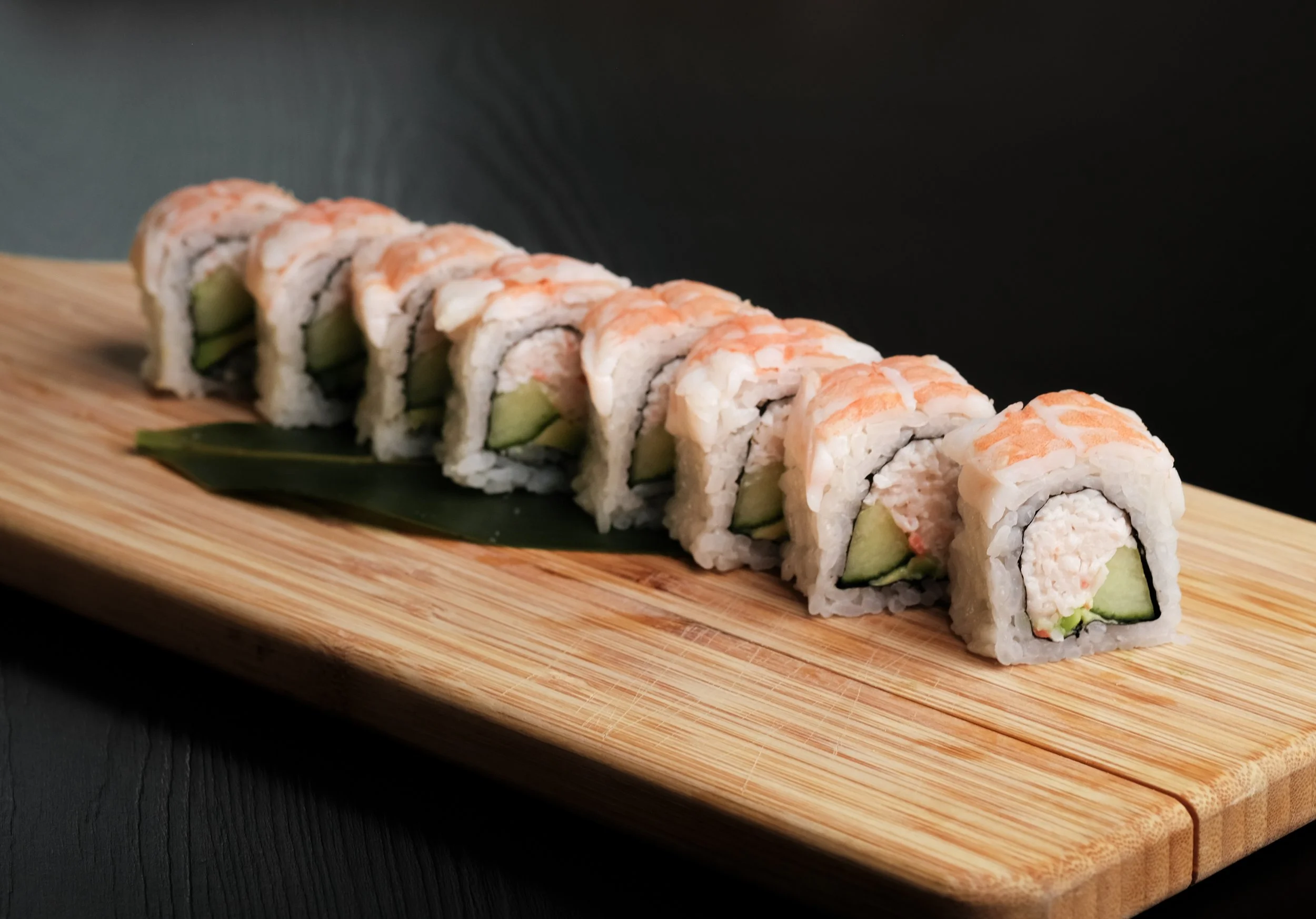 A row of sushi rolls with shrimp on top, containing cucumber and crab, placed on a wooden serving board with a green leaf underneath.