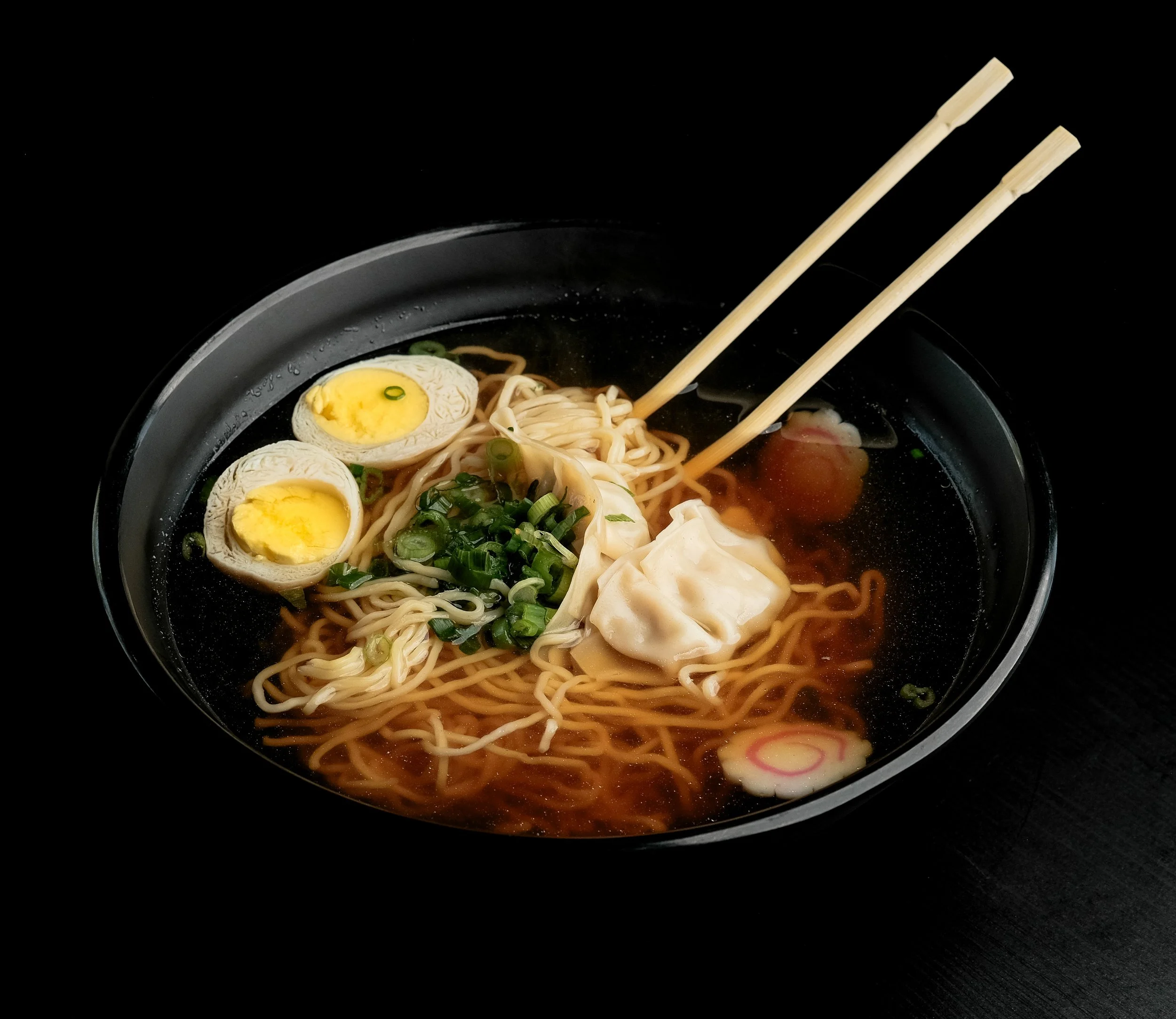 Bowl of ramen with soft boiled eggs, chopped green onions, dumplings, and narutomaki with chopsticks resting on the side.