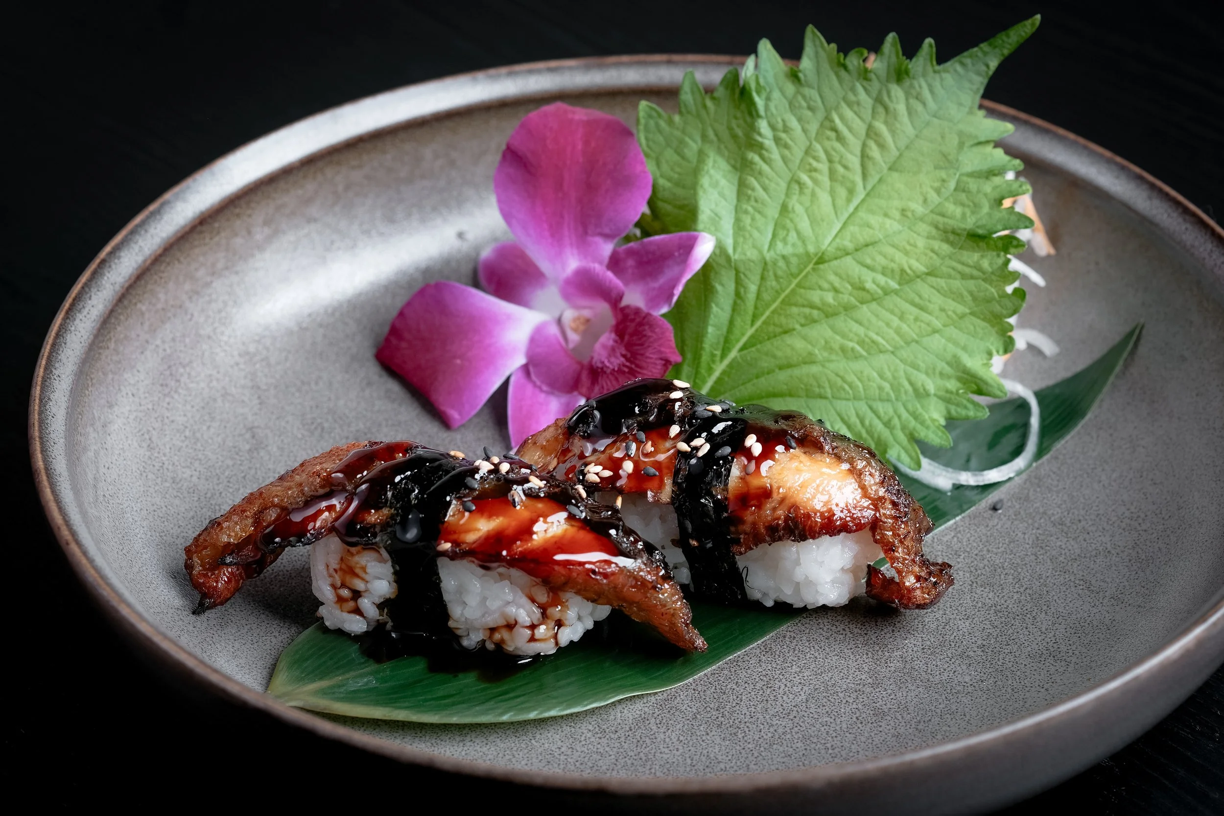 Two pieces of sushi topped with eel sauce and sesame seeds, garnished with a green shiso leaf, a purple orchid flower, and thinly sliced white onion on a gray ceramic plate.