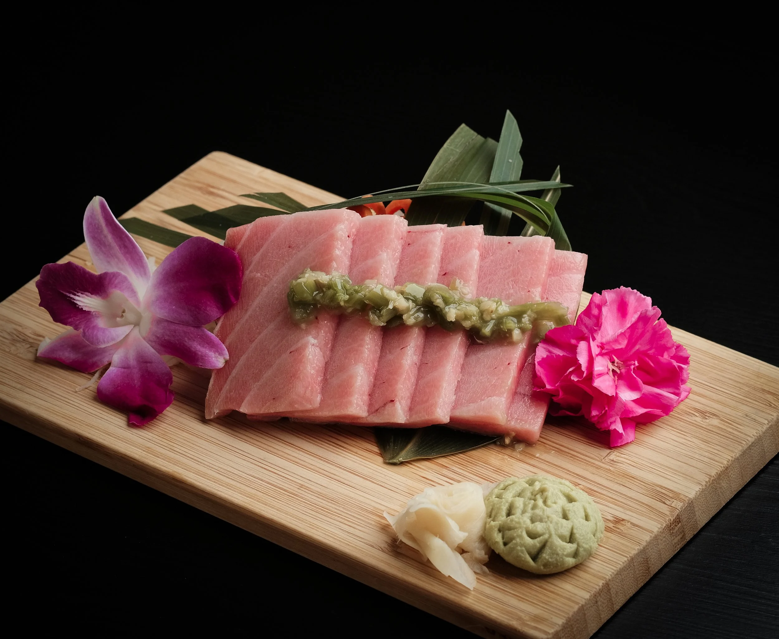 Slices of raw fish, garnished with green sauce, green leaves, orchid flowers, wasabi, and pickled ginger on a wooden serving board.