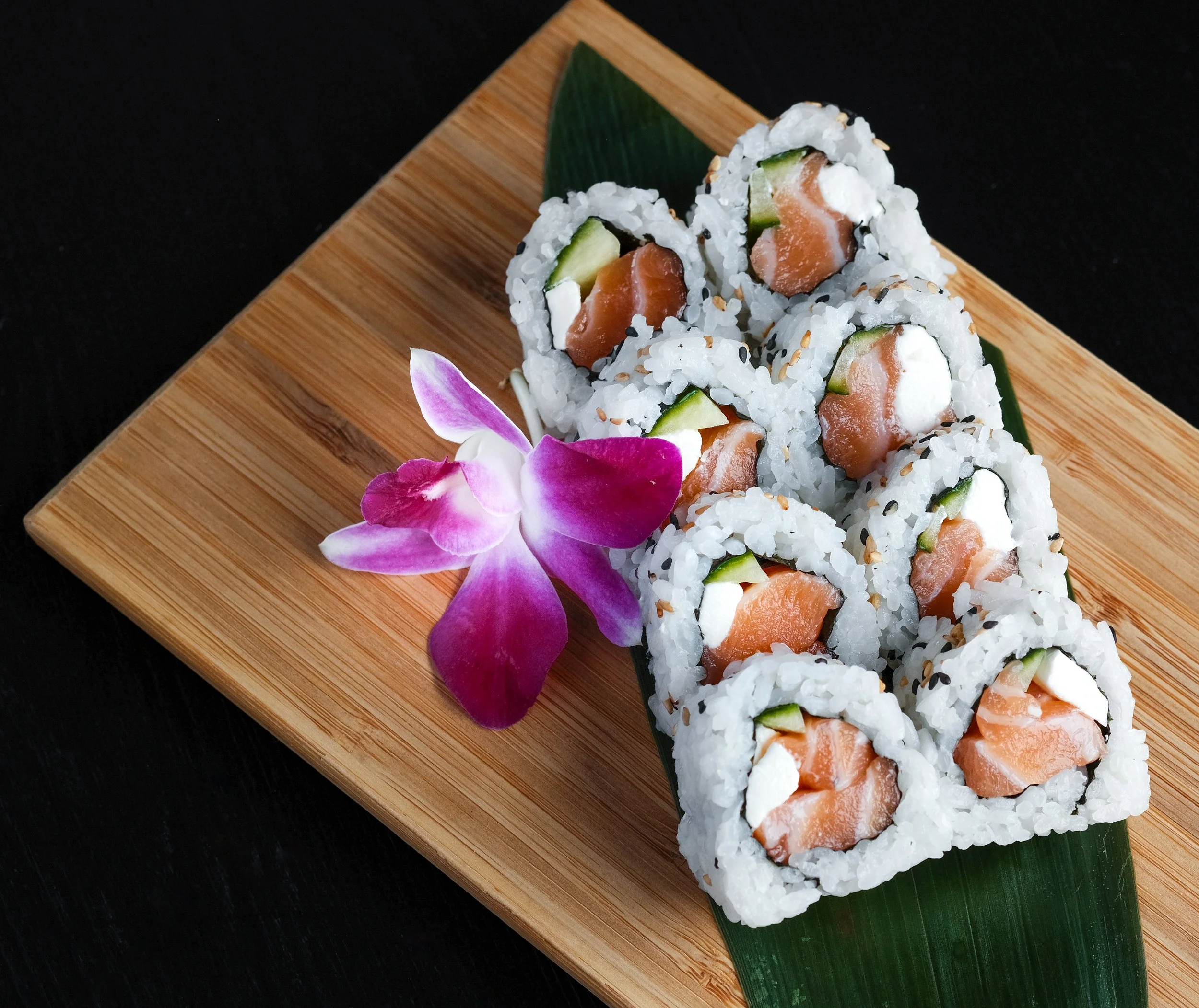A wooden tray with six sushi rolls containing salmon, cream cheese, and cucumber, garnished with sesame seeds, a green leaf, and a purple orchid flower.