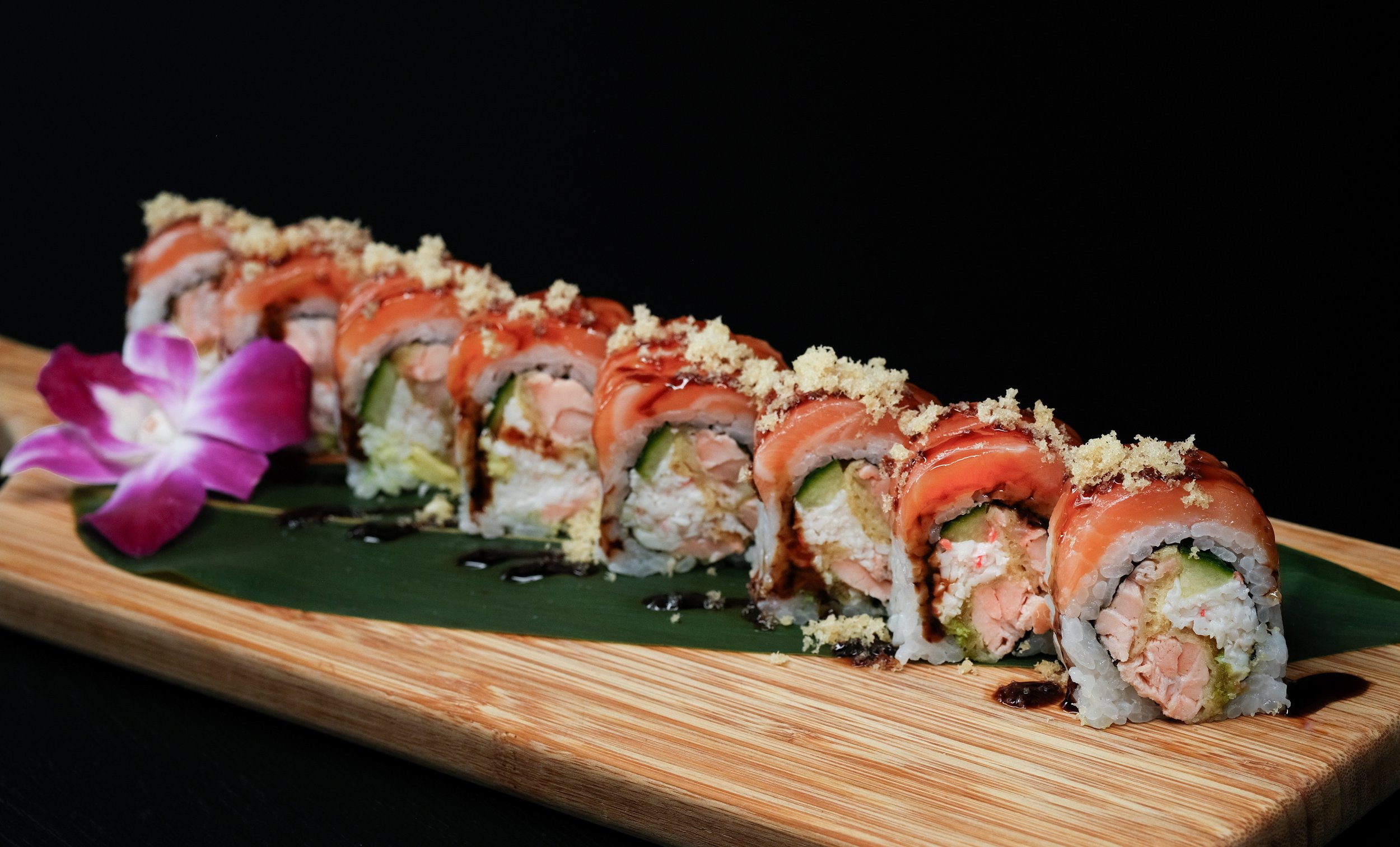 Slice of sushi roll with seafood, cucumber, rice, wrapped in salmon, topped with sauce and crumbs, garnished with a pink orchid flower on a wooden platter with green leaf