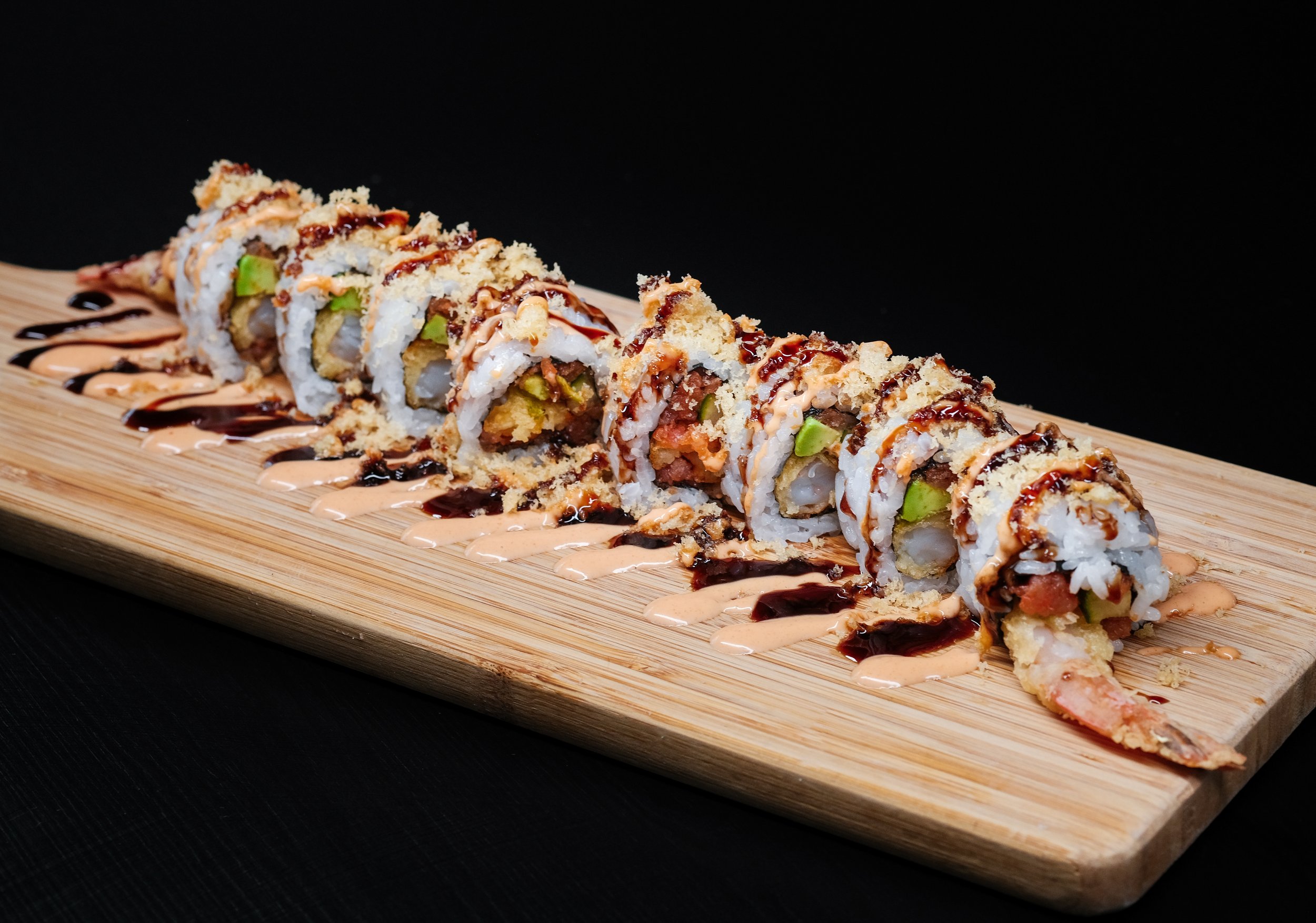 Sushi roll with shrimp tempura, avocado, and crab, topped with drizzles of spicy mayo, eel sauce, and crispy tempura flakes, served on a wooden plate with decorative sauces.