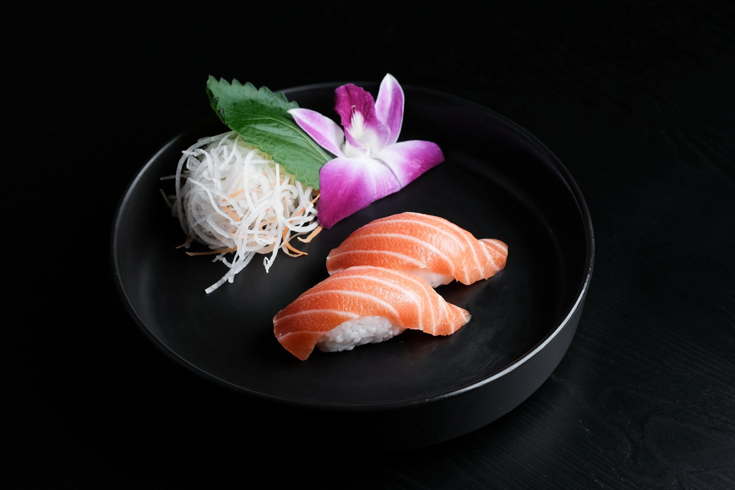 Two pieces of salmon nigiri sushi arranged on a black plate, garnished with shredded daikon and carrot, an edible purple and white orchid flower, and a green leaf, set against a black background.
