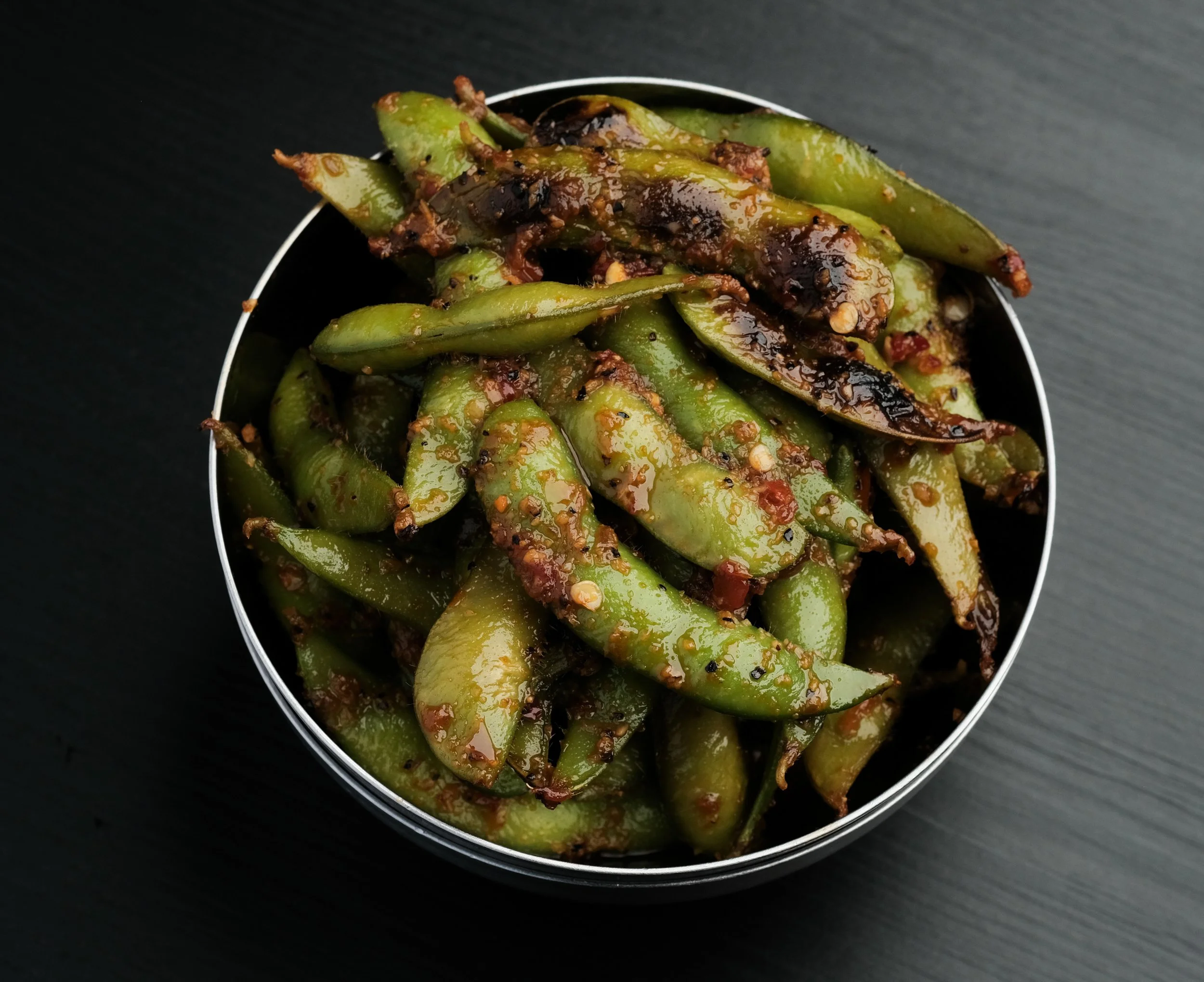 A bowl of cooked edamame beans with dark sauce and spices on a dark surface.
