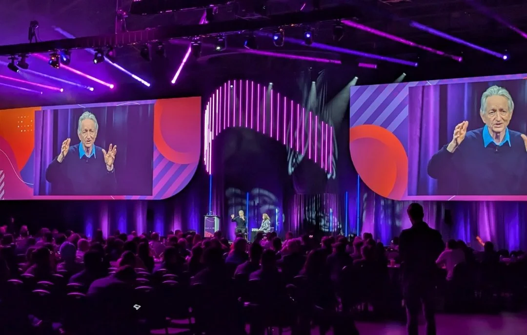 A photo of Dr. Geoffrey Hinton delivering the keynote presentation at OCI's DiscoveryX Conference in Toronto, April 30,2026. Stage is light with brand colour gradient of purple, pink and orange, with stage lighting, Hinton on screen gesturing.