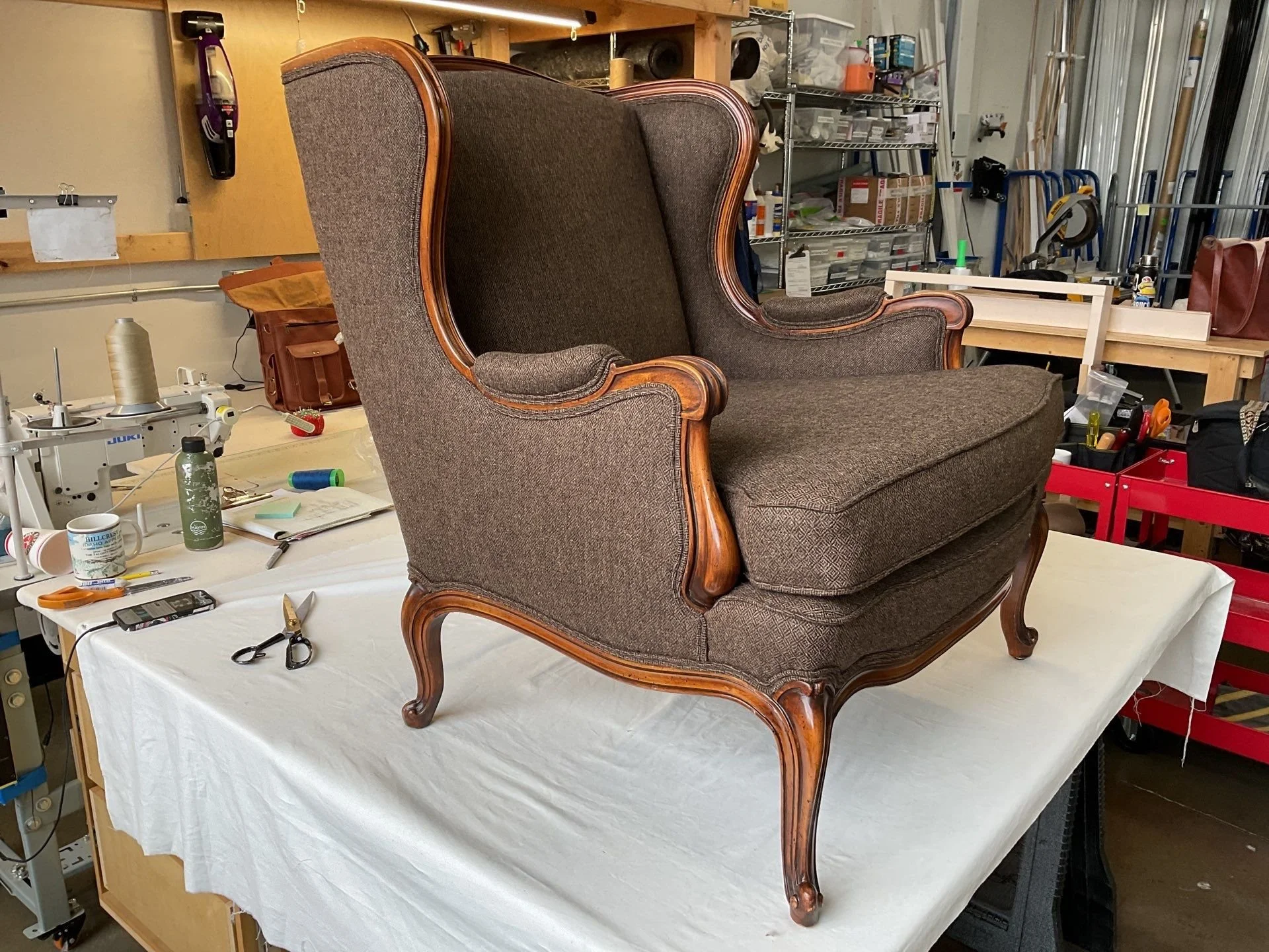 An antique wooden armchair with dark upholstery placed on a table covered with white cloth in a workshop or storage room with various tools and shelves .Created by LP Studio in Woodinville, WA.