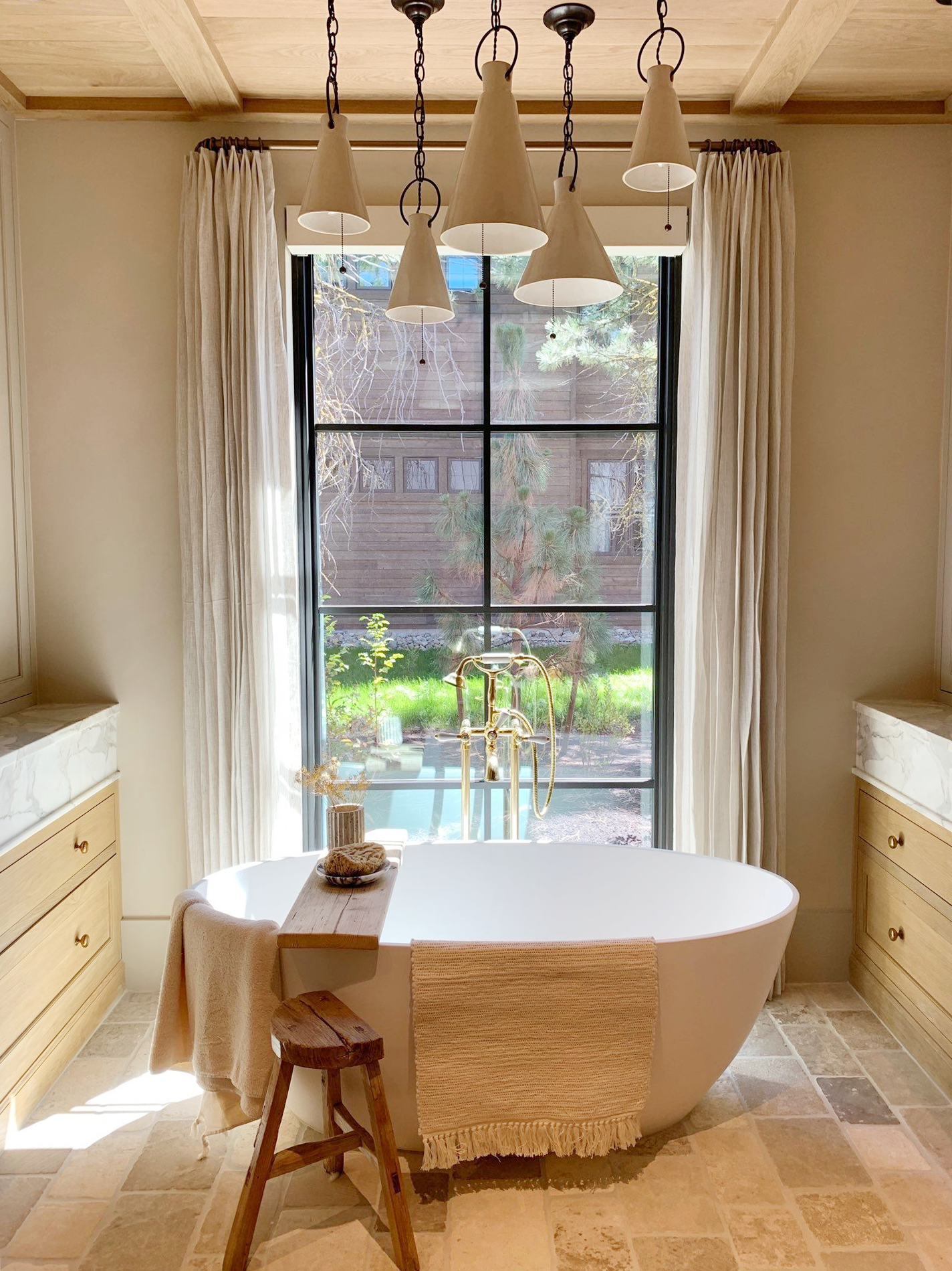 A bathroom with a freestanding bathtub placed in front of a large window, which lets in natural light and shows outdoor trees and a wooden fence. There are cream curtains on the window - Created by LP Studio in Woodinville, WA.