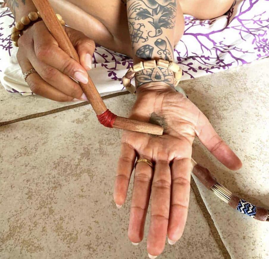 Person holding a wooden pipe and a small amount of kambo in hand, with tattoos visible on their arm.