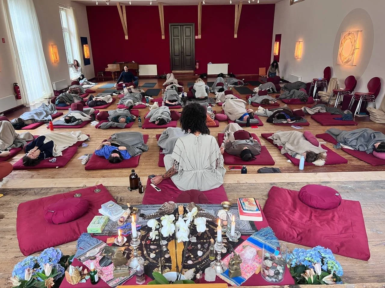 Meditation session in a large room with people lying on mats and cushions. An instructor sits at the front, facing the participants who are in peaceful postures. The room has wooden floors, red and white walls, and ambient lighting with some decorative elements.
