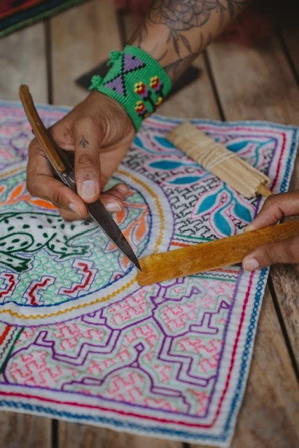 Person working on a colorful, outlined embroidery or textile design with a bright green beaded bracelet and wooden tools.