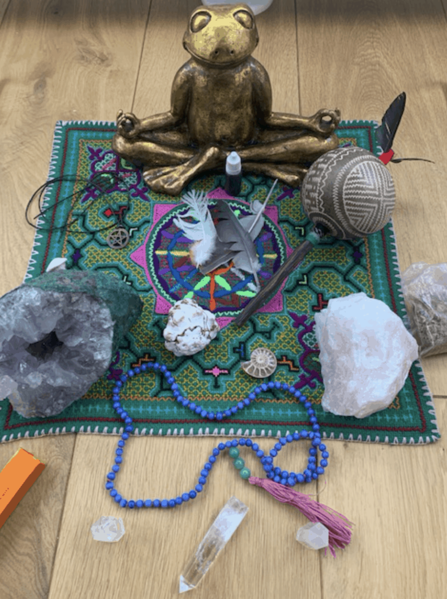 Meditation altar with a brass frog statue, colorful mat, crystals, feathers, beads, and a tribal pattern rattle.