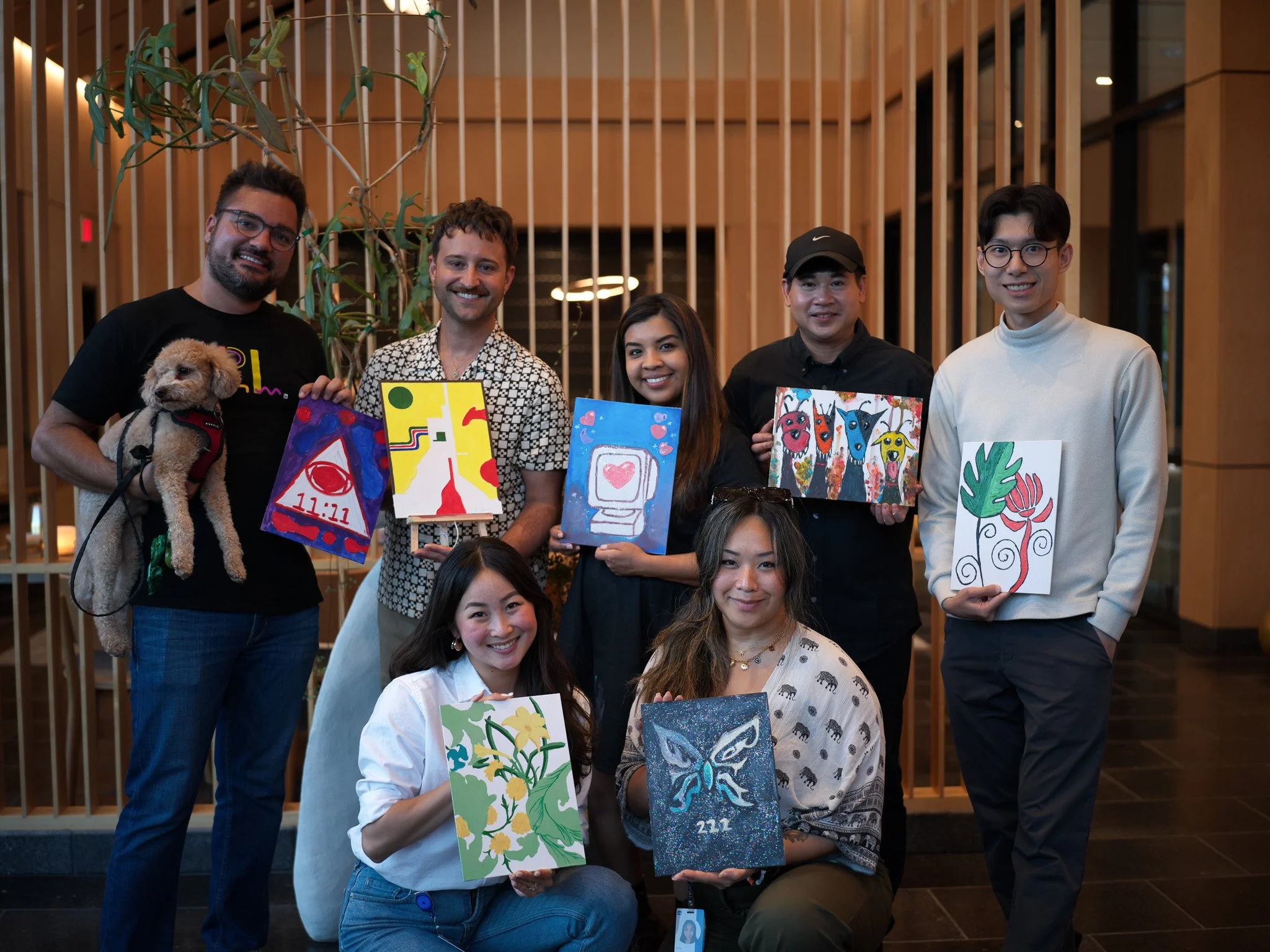 Group of eight people, five standing and three kneeling, holding colorful paintings, inside a modern building with wooden panels and plants.