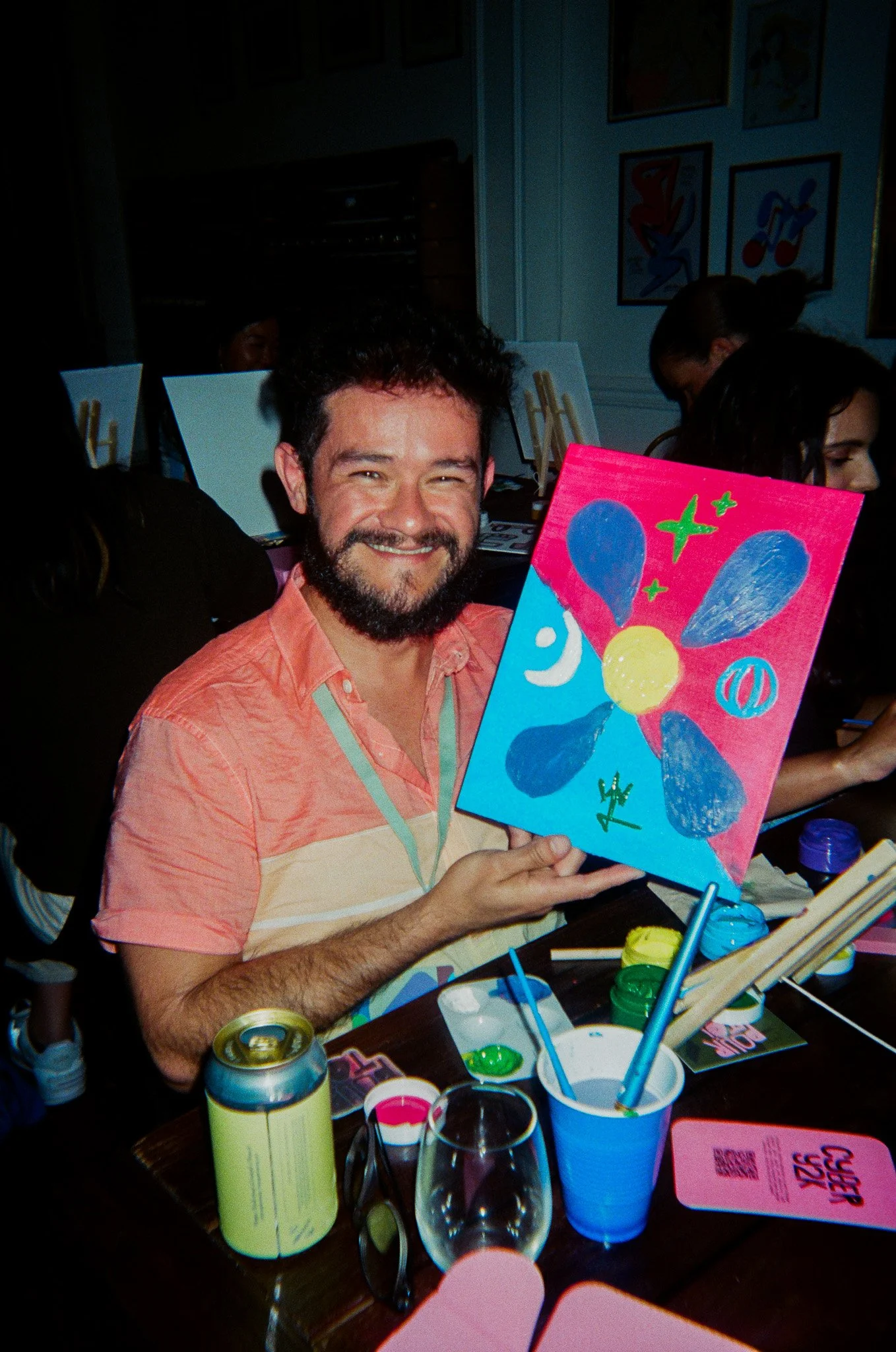 A man smiles while holding a colorful painting at a Pour Toi paint n sip event. The table in front has drinks and art supplies.