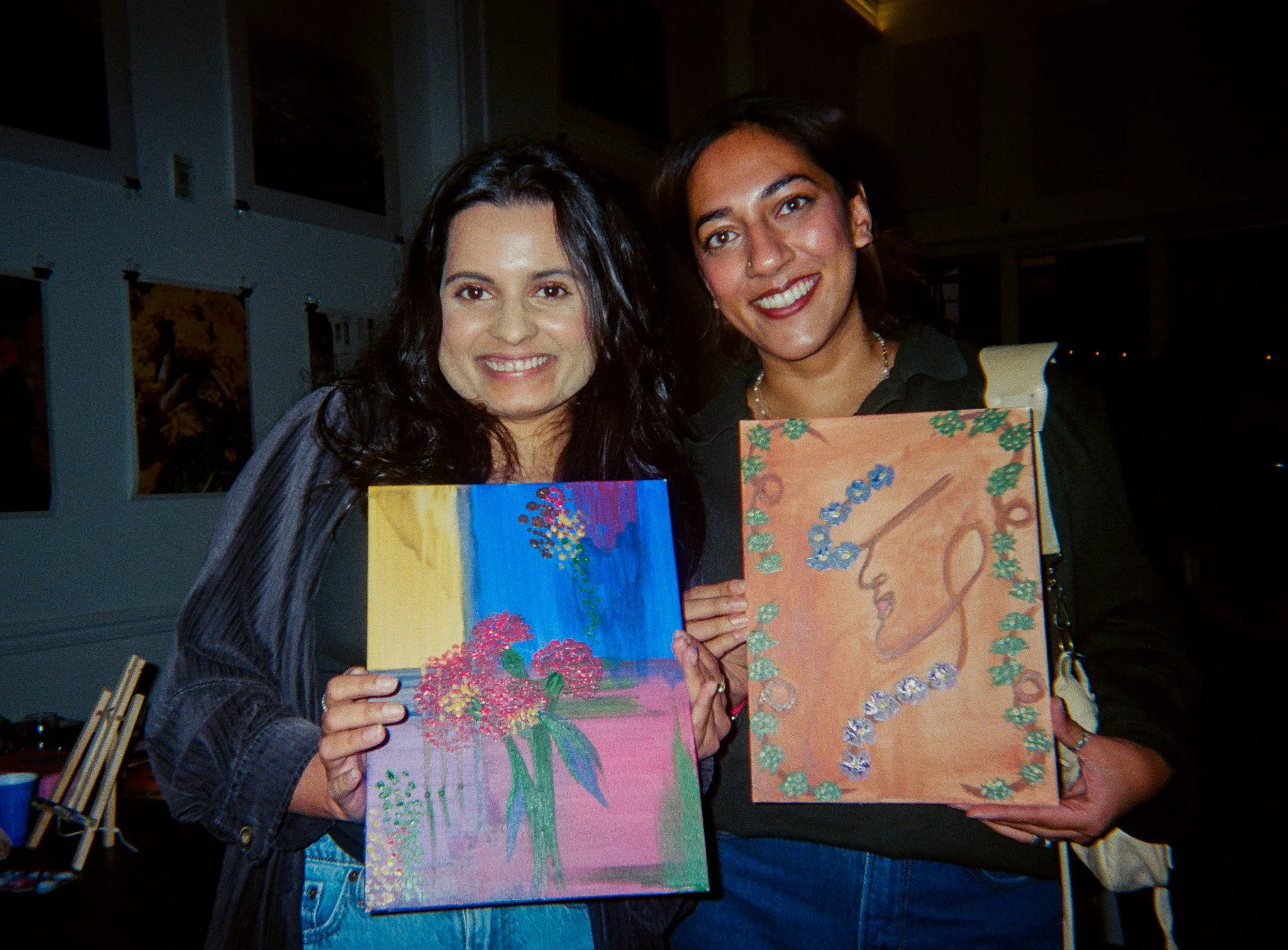 Two women smiling and holding colorful paintings at a Pour Toi paint n sip event.