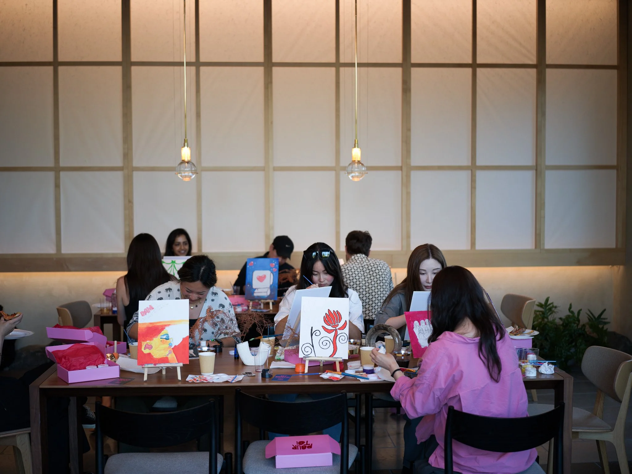 Group of women and men sitting at a table, painting and creating art projects inside a well-lit room with wooden panels and hanging pendant lights.
