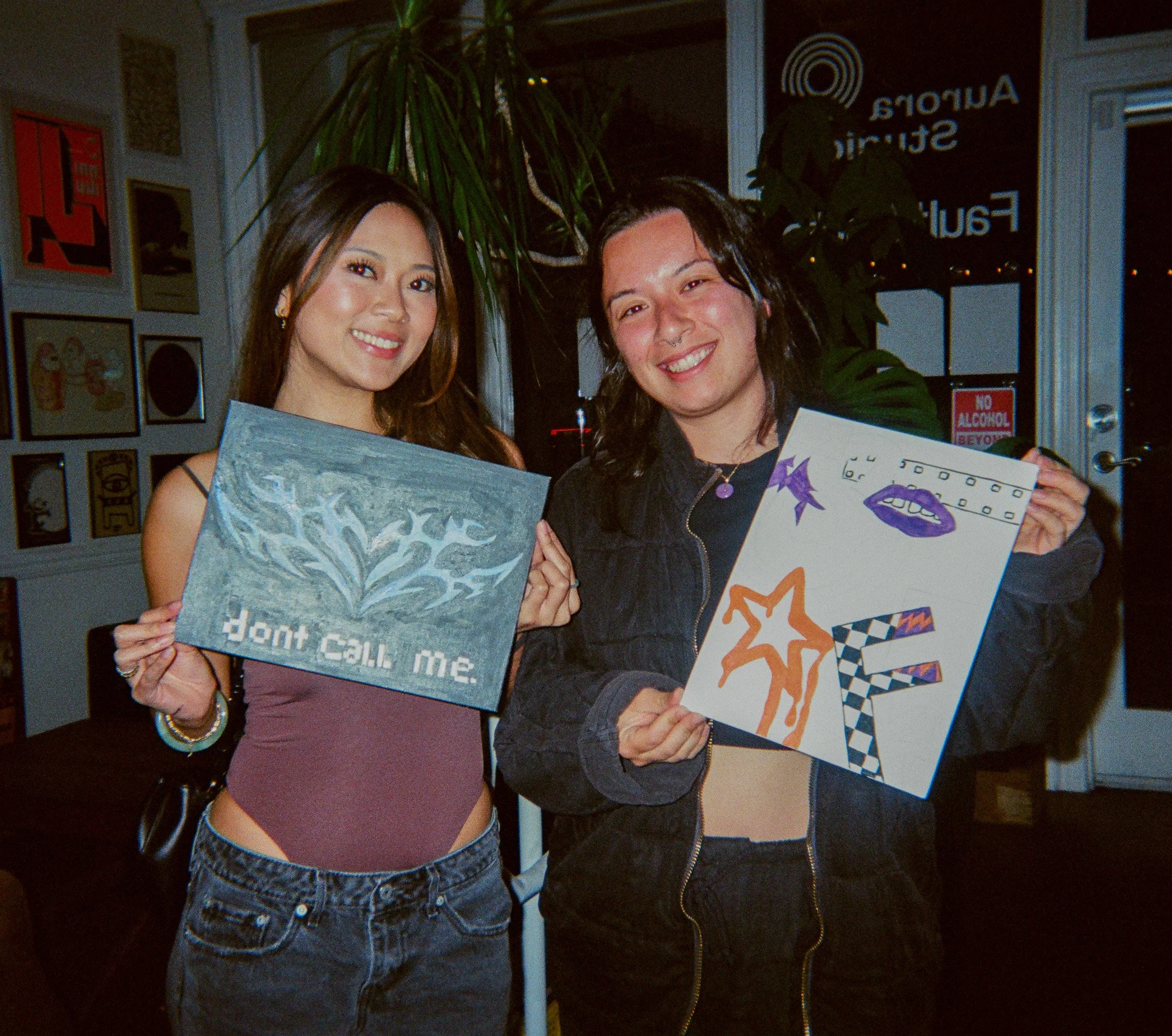 Two young women smiling and holding colorful art pieces at a Pour Toi paint n sip event.