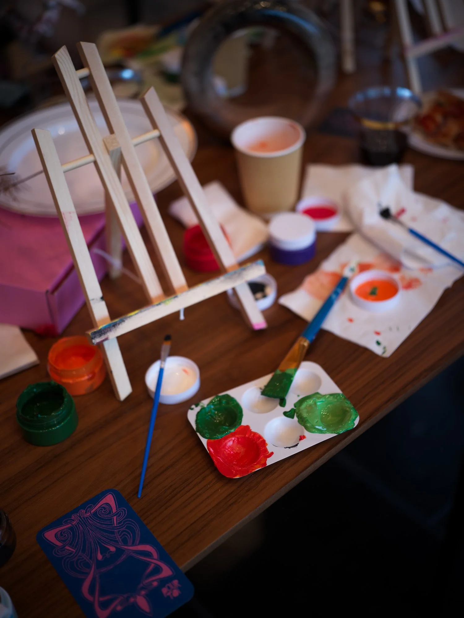 A wooden mini easel on a table surrounded by paint bottles, brushes, and paper with painted orange and green designs, indicating a watercolor painting activity.