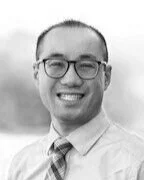 A smiling man wearing glasses, a collared shirt, and a tie, standing outdoors.