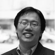 Black and white photo of a smiling man with glasses and straight hair, in an outdoor setting.