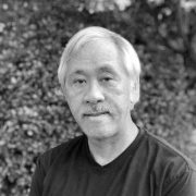 Black and white photo of a man with gray hair and a beard, wearing a dark shirt, standing outdoors with trees in the background.
