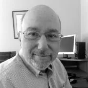 A middle-aged man with glasses and a beard smiling in an office space with a computer and framed picture in the background.