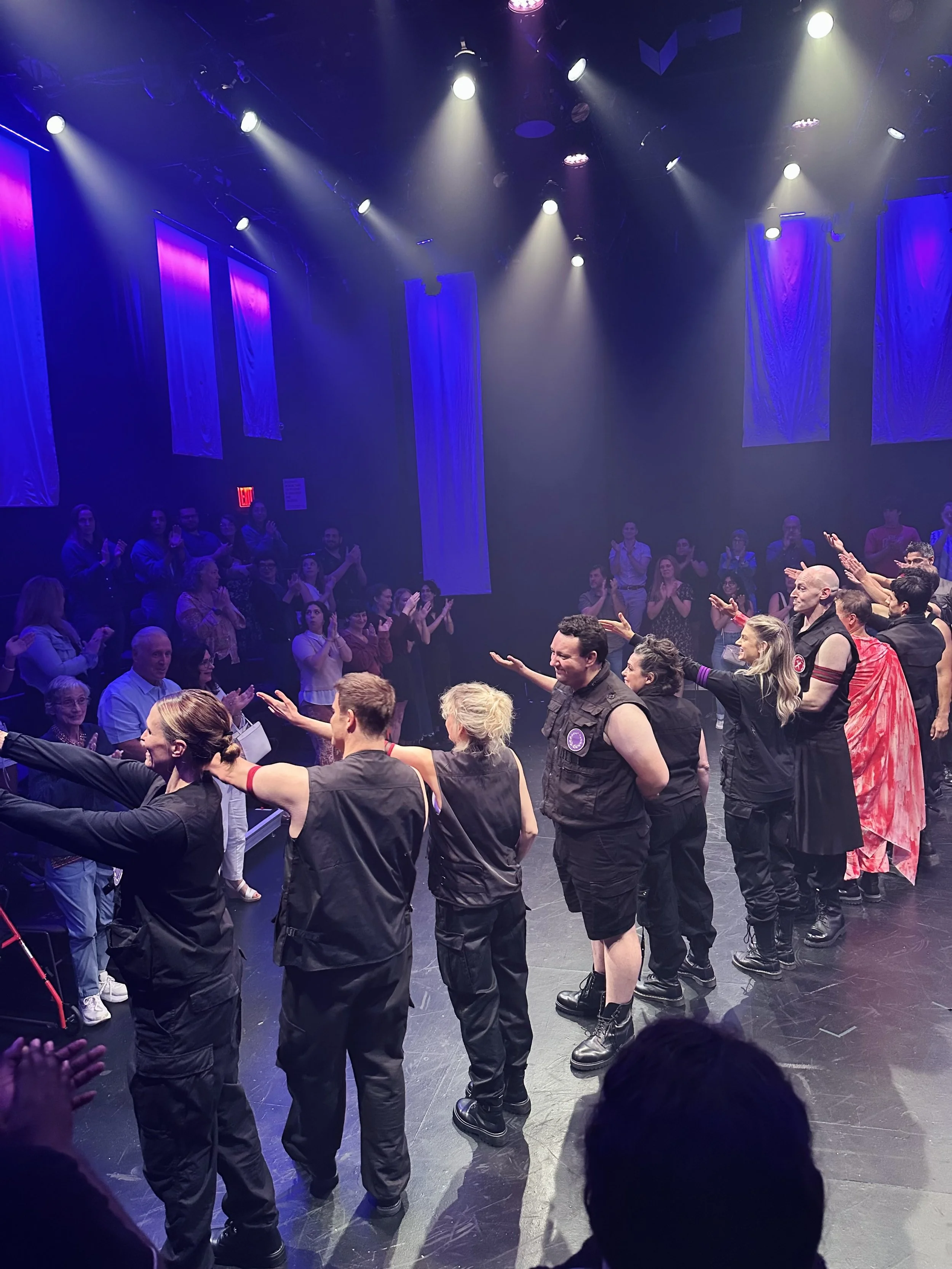 A wide view of an ensemble cast performing a scene on an intimate stage under striking purple and blue spotlights. The actors are wearing dark, modern costumes, with the audience seated closely around the performance space.
