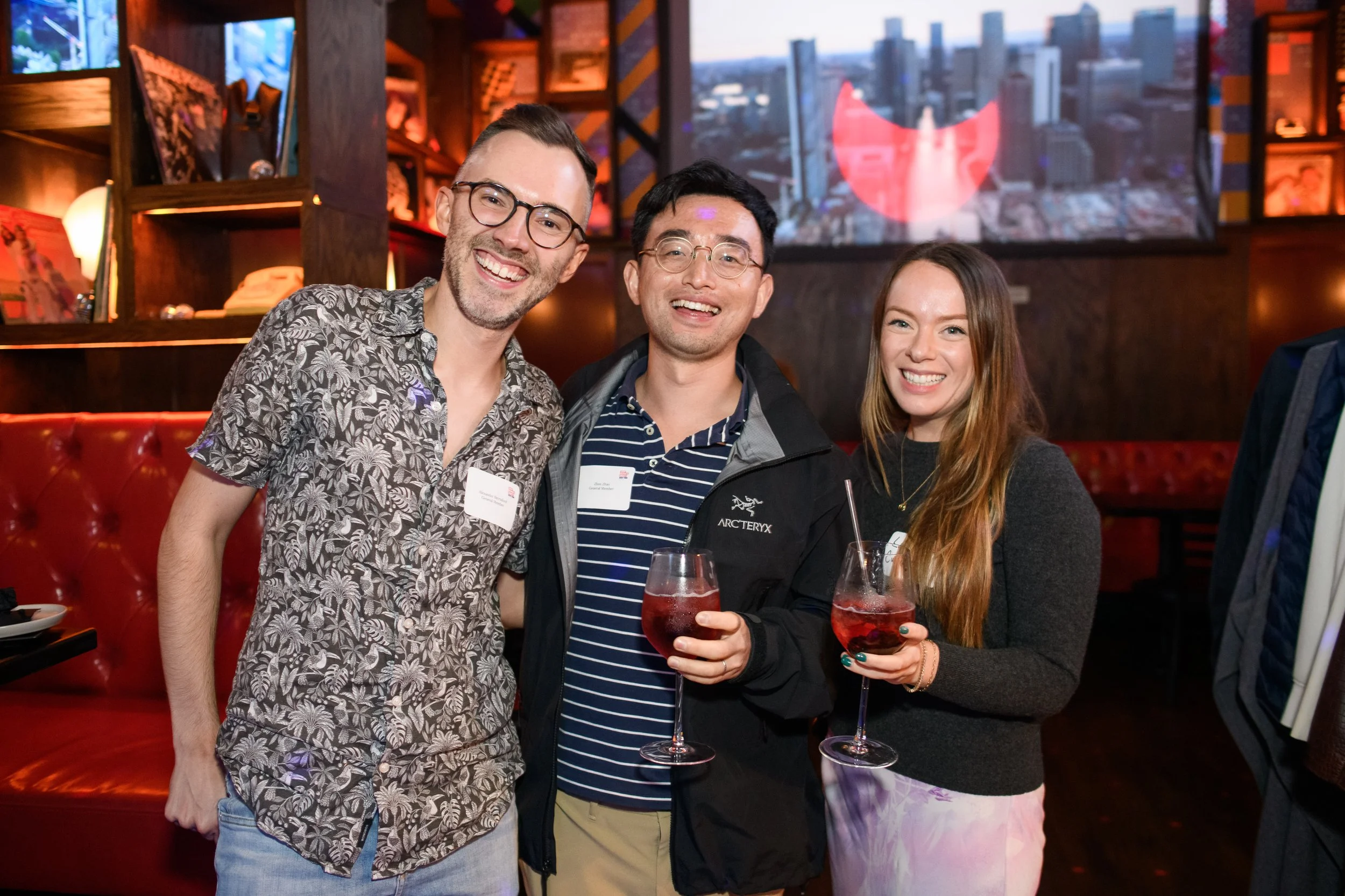 Three people with cocktails at a dimly lit bar, smiling at the camera.