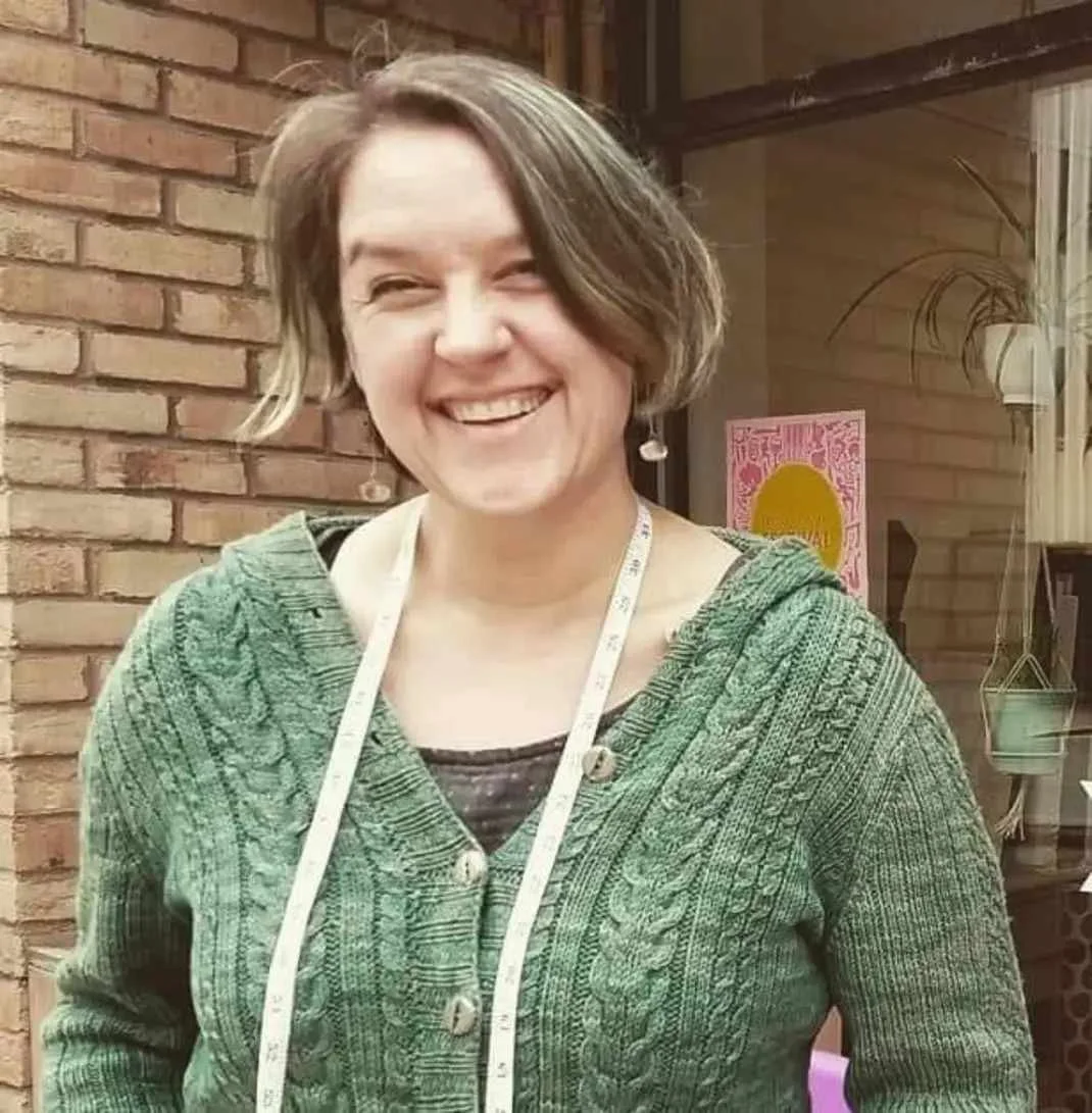 Person smiling in a green sweater with a measuring tape around their neck
