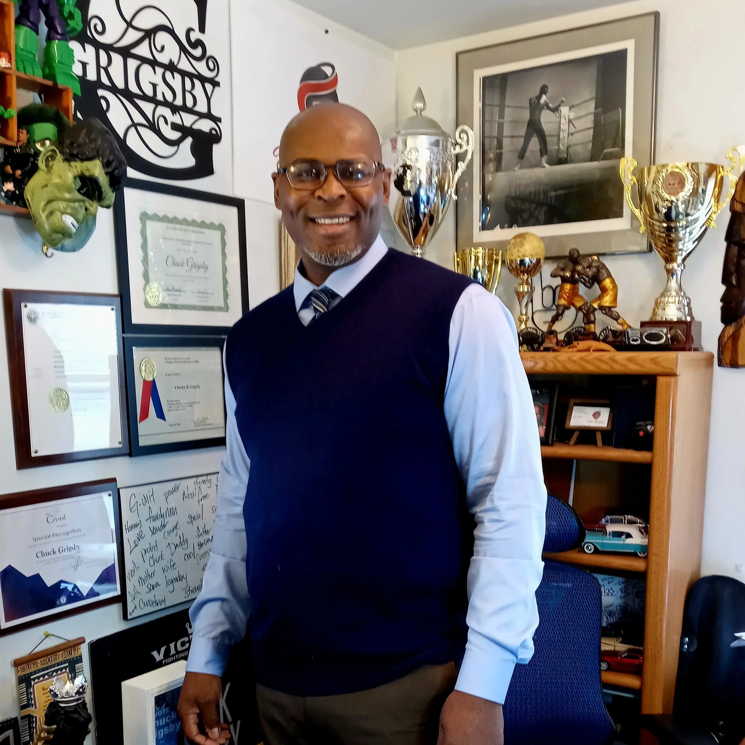 A person in a sweater vest and tie standing smiling in front of a wall of picture frames