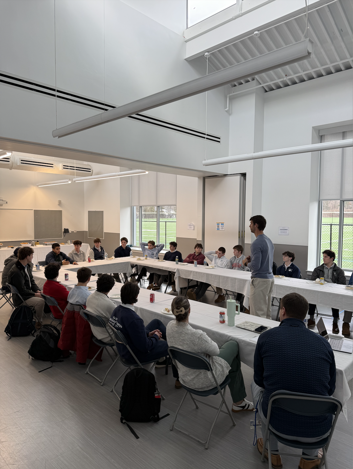 A man standing and speaking to a group of young men seated at a U-shaped table in a bright, modern room with large windows and white walls.