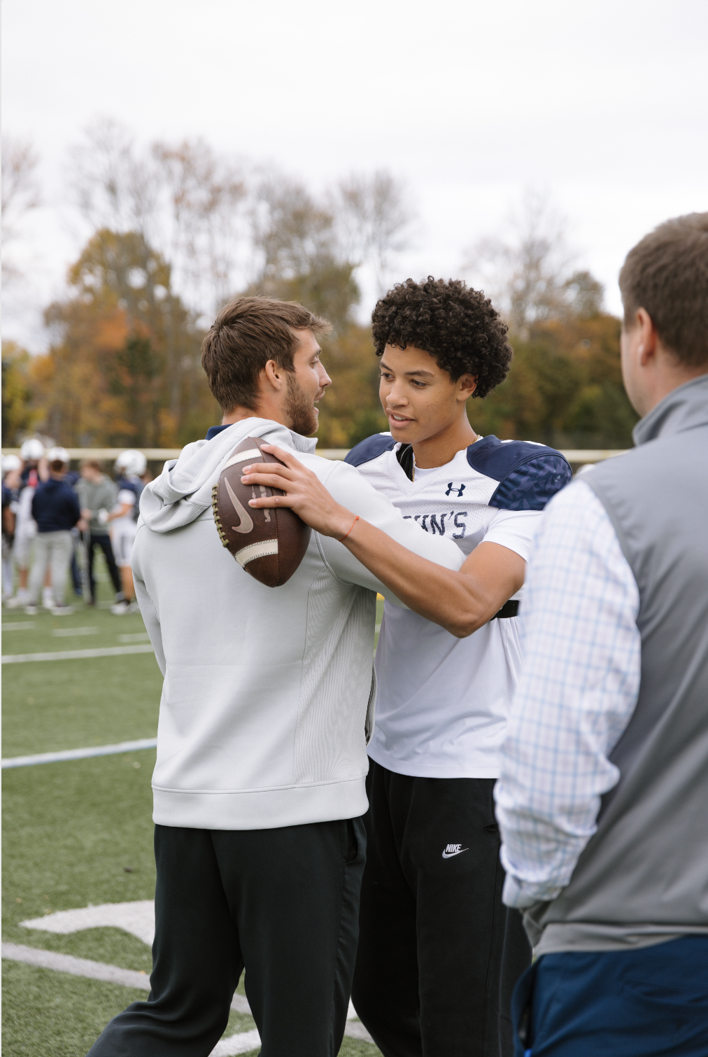 Two young men on a football field, one in a white hoodie holding a football and the other in a football uniform, are interacting with each other while a third person stands nearby. In the background, a group of people are gathered on the sideline, wi