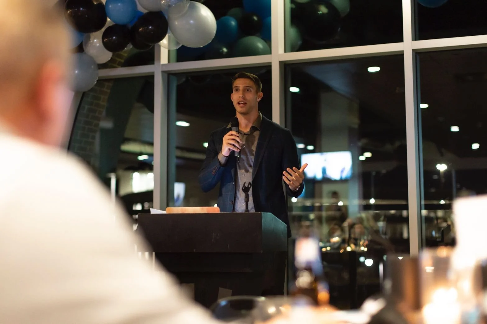 A young man in a dark blazer and gray shirt is speaking into a microphone at a table during a public event or speech, in a room decorated with balloons in the background.