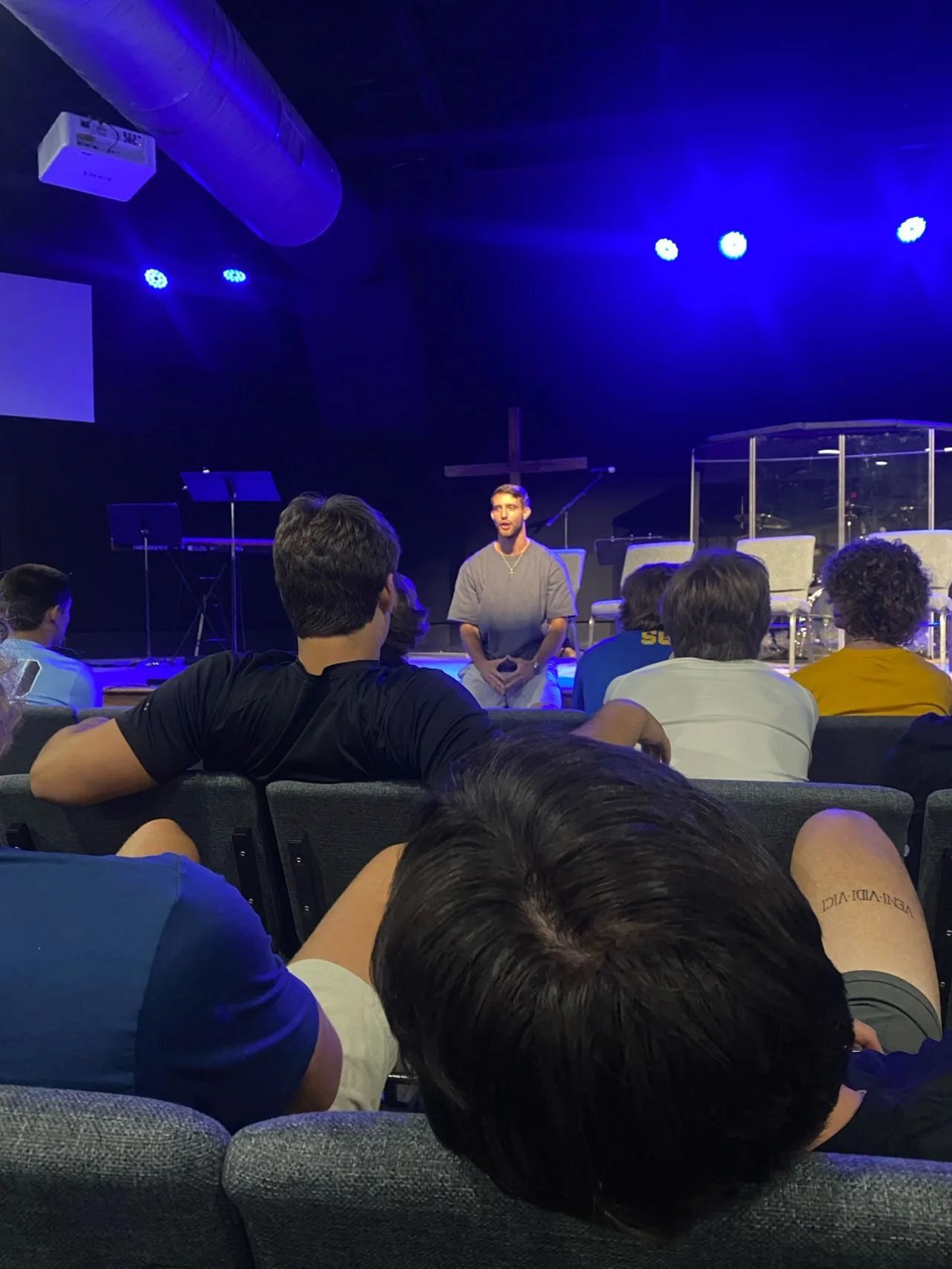 A man speaking in front of an audience in a church setting with a cross and a stage, illuminated by blue stage lights.