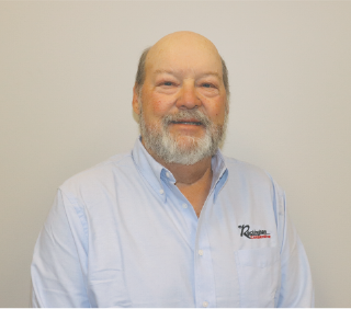 A smiling middle-aged man with a beard and mustache, wearing a light blue button-up shirt with a company logo, standing against a plain background.