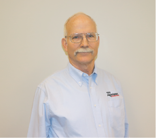 An older man with glasses and a mustache wearing a light blue button-up shirt standing against a plain background.
