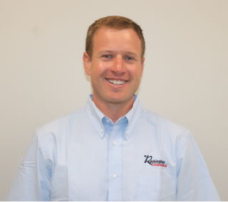 A man with short light brown hair, wearing a light blue collared shirt with a logo, smiling against a plain white background.