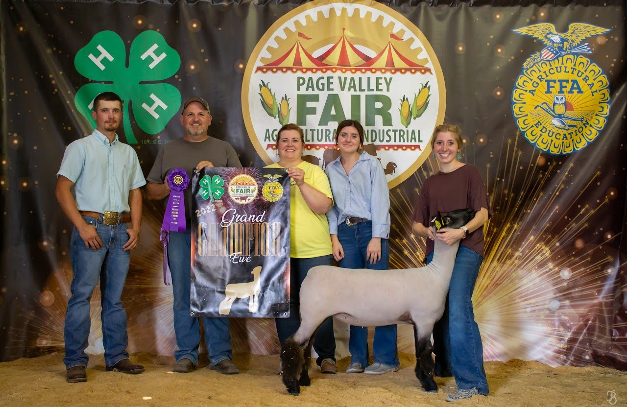 Group of five people standing indoors at the Page Valley Fair, with a backdrop featuring the fair's logo and symbols of agriculture. One person holds a large purple ribbon, another holds a banner reading 'Grand Champion Eve,' and a young woman on the far right holds a goat, with a white goat standing in front of her. The group appears to be celebrating a livestock competition victory.