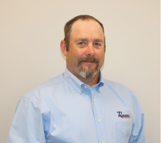 A man with short brown hair, a beard, and mustache wearing a light blue collared shirt with a company logo on the pocket, standing against a plain white background.