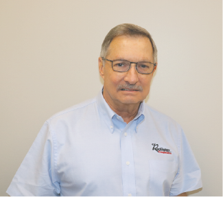 A middle-aged man with glasses and a mustache, wearing a light blue button-up shirt with a logo, standing against a plain light-colored wall.