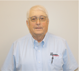 Elderly man with white hair and glasses wearing a light blue shirt with a company logo, standing against a plain background.