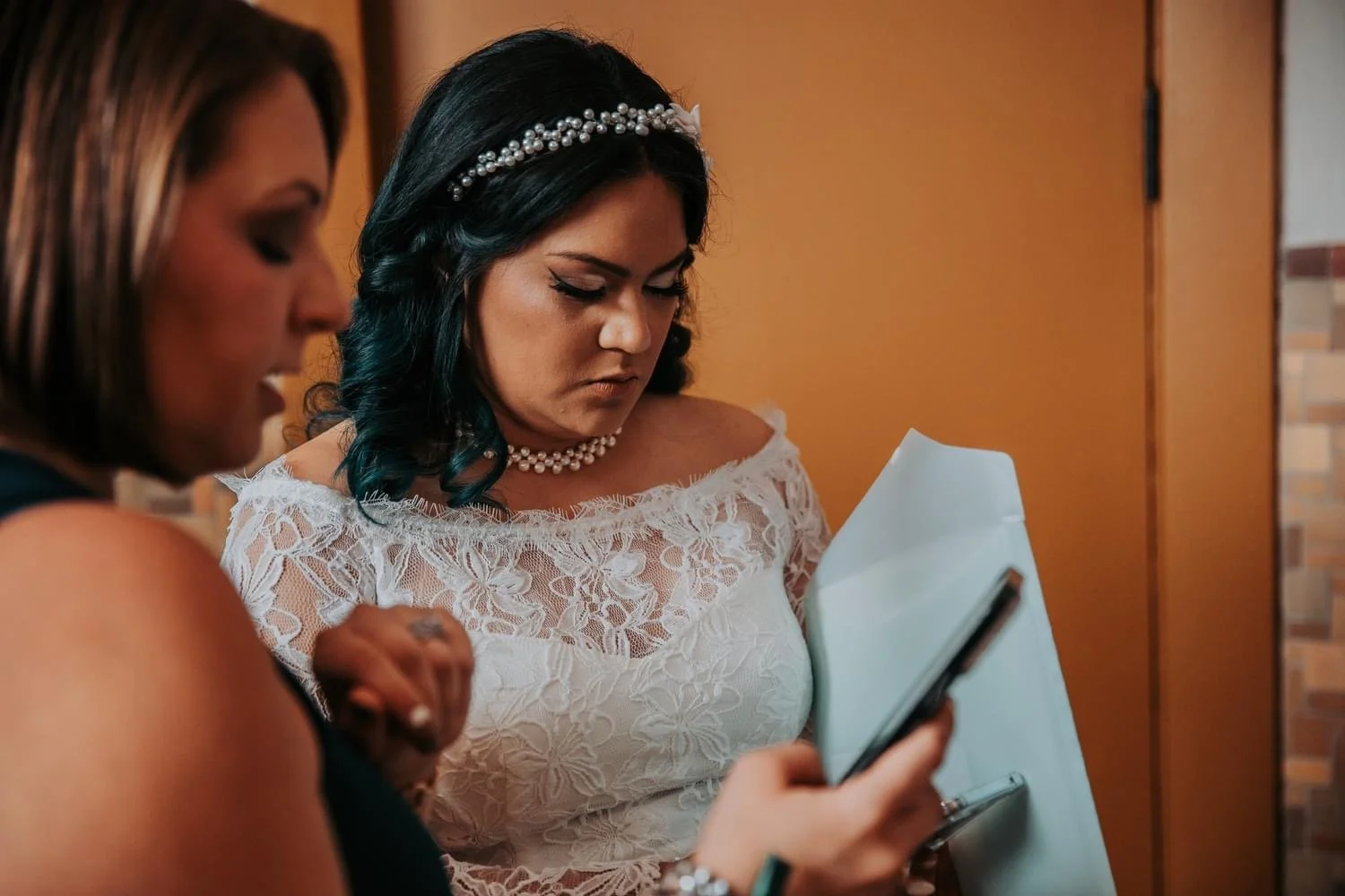 Woman in lace dress with pearl accessories looking at phone, another person nearby.